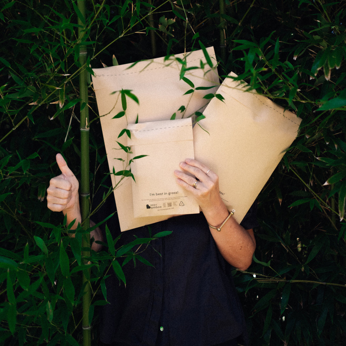 A person holding three Better Packaging brown bamboo envelopes in front of bamboo shoots
